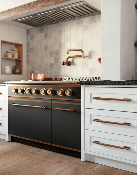 Close-up of a black luxury range with copper and brass accents and hardware, a copper pot filler faucet, and a stainless steel range hood, flanked by white shaker cabinets with long copper pulls, all set above a light wood floor and against a light grey patterned tile backsplash.