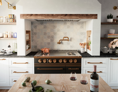 Traditional-style kitchen featuring a black and brass luxury range by La Cornue or similar, set within a stucco and rough wooden beam alcove, with a brass pot filler faucet, grey patterned tile backsplash, and white lower cabinets with copper hardware, and a light wood island in the foreground.