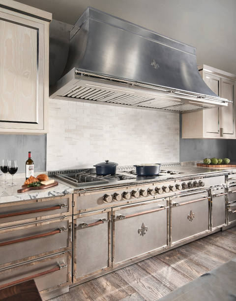 Luxury kitchen with a large brushed stainless steel range hood and a long L'Atelier Paris-style stainless steel range with brass and wood handles and Fleur de Lis accents, set against a white marble subway tile backsplash and light wood grain upper cabinets.