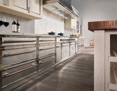 Low-angle view of a luxury modern kitchen featuring a long bank of stainless steel base cabinets and a range by L'Atelier Paris or similar high-end brand, with wooden handles and polished chrome trim, set against a white subway tile backsplash, with a butcher block cutting board visible on the right.