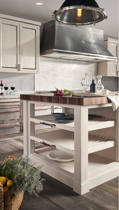 Vertical view of a rustic-chic kitchen island featuring a thick dark wood butcher block countertop and white slatted open shelving, with a basket of herbs on the dark wood floor. In the background, a stainless steel range hood with a Fleur de Lis hangs over a white subway tile backsplash and stainless steel range with copper handles.