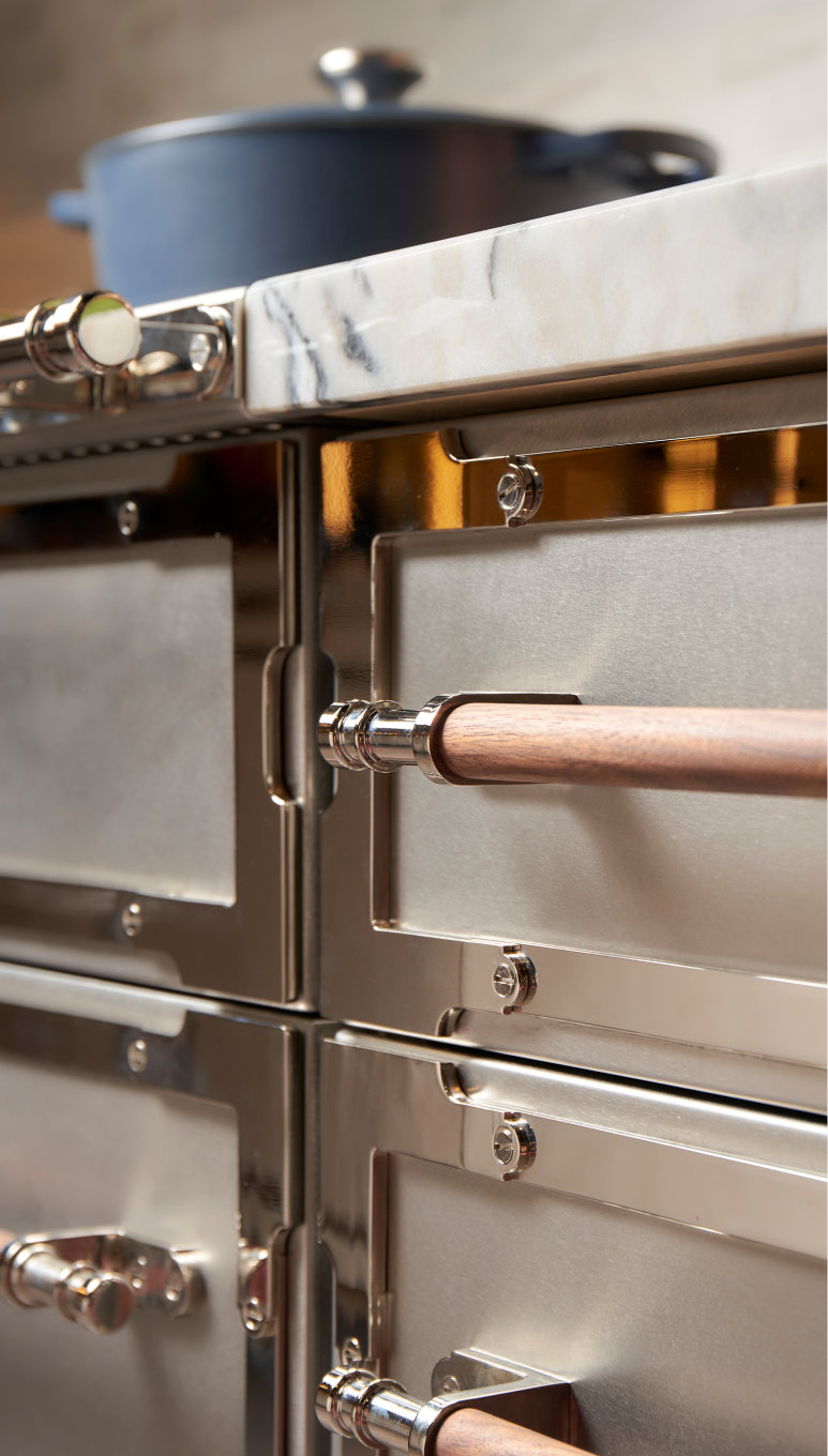 Close-up detail of a luxury stainless steel range or oven column featuring polished chrome trim and wooden horizontal handles, topped with a white and grey veined marble countertop and a blue cast iron pot in the background.