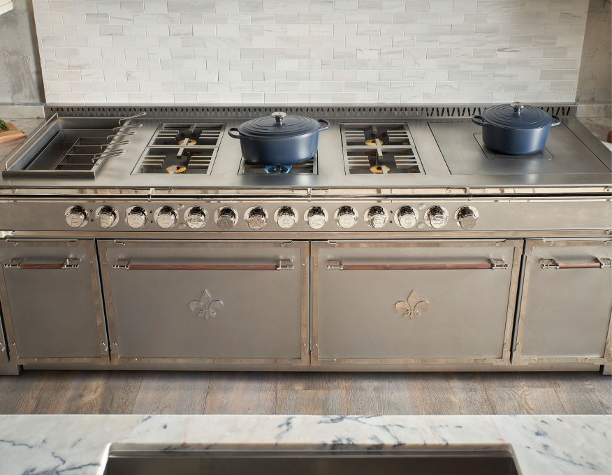 Close-up of a large stainless steel luxury range or cooktop by a brand like L'Atelier Paris, featuring brass and wood handles, chrome control knobs, and Fleur de Lis accents on the oven doors, with blue cast iron pots on the burners and a marble subway tile backsplash.