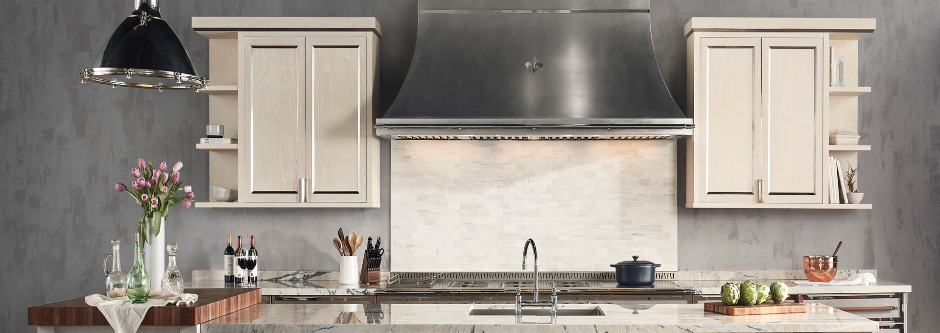 Luxury kitchen with a large stainless steel range hood featuring a Fleur de Lis accent, mounted over a white iridescent subway tile backsplash and a stainless steel La Cornue range and sink, all set beneath light cream cabinets against a dark gray textured wall.