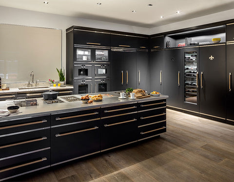 A professional wide shot of a high-end, contemporary kitchen featuring a massive matte black island with a stainless steel cooktop. The black cabinetry and island drawers are strikingly accented with brass trim and long wooden handles. The back wall includes a bank of integrated black ovens, refrigerators, and a glass-door wine refrigerator with a subtle Fleur-de-lis emblem on the door panel. The kitchen is completed with warm wood flooring.