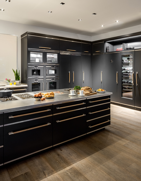 A wide shot of a luxurious, modern black kitchen featuring an impressive central island. The matte black cabinetry is accented with gold-tone or brass trim and wooden drawer pulls. The island has a professional-grade stainless steel cooktop and counter space, with breakfast items arranged on top. The back wall features a bank of built-in black appliances, including multiple ovens, integrated refrigerators, and a large glass-door wine refrigerator. The kitchen has recessed lighting and dark wood flooring.
