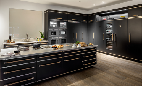 Modern luxury kitchen featuring a massive matte black island with a built-in cooktop, accented by unique brass trim and long wooden handles, and a wall of integrated appliances including a glass-door wine fridge.