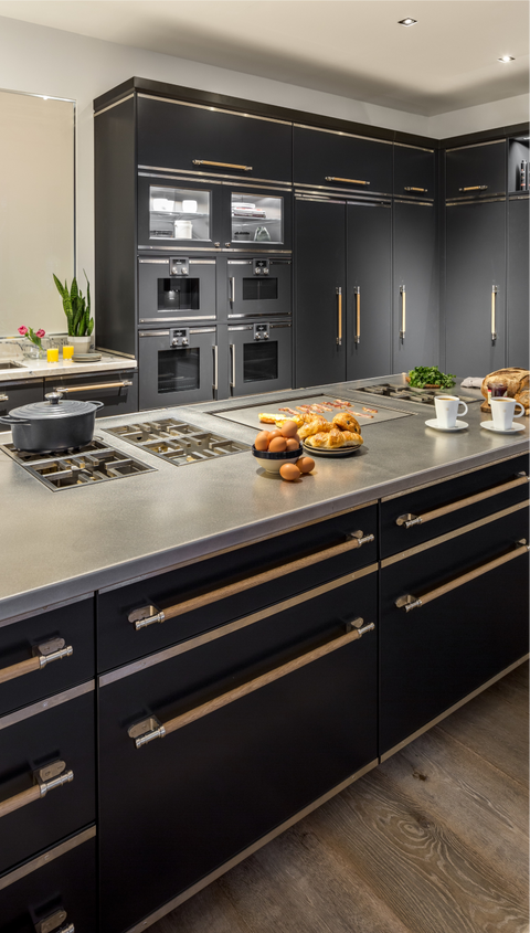 A detailed vertical close-up of a luxury kitchen island featuring a matte black base and a seamless stainless steel cooktop with multiple gas burners. The drawers are accented with brass trim and wooden horizontal pulls. The background shows a bank of integrated black appliances, including ovens and refrigeration units, also featuring the brass and wooden hardware. Breakfast items are casually placed on the island counter.