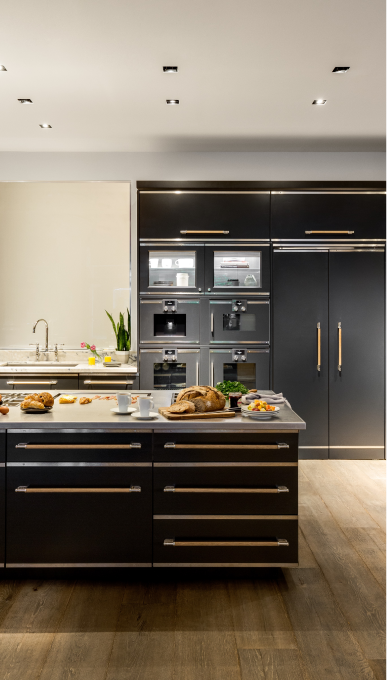 A vertical shot of a modern black kitchen island with a stainless steel countertop and breakfast items displayed. The matte black drawers feature brass trim and long wooden pulls. Behind the island is a wall of integrated black appliances, including multiple ovens and a refrigerator, with stainless steel and wooden accents. The kitchen has warm wood flooring and recessed ceiling lights.