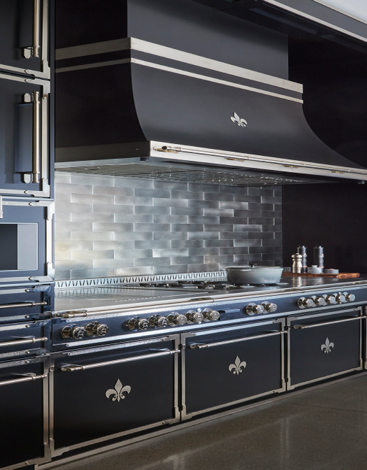 Vertical view of a high-end black La Cornue range and custom black canopy hood with stainless steel trim and a Fleur de Lis accent, set against a metallic silver subway tile backsplash. The range features chrome control knobs and Fleur de Lis oven door emblems, showcasing a luxurious, modern design.