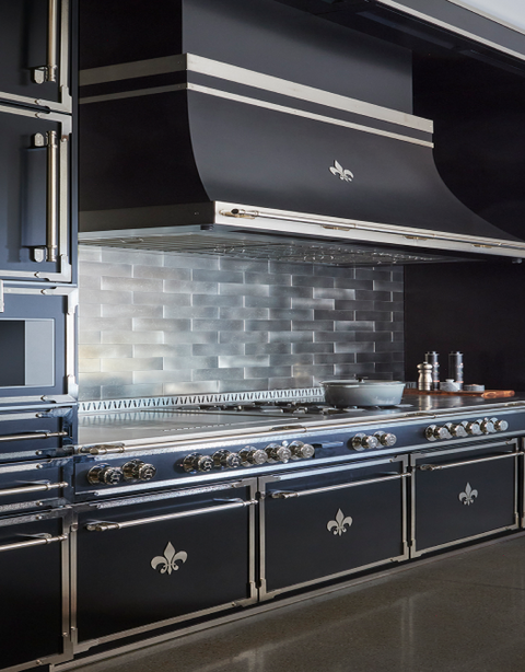 Vertical view of a high-end black La Cornue range and custom black canopy hood with stainless steel trim and a Fleur de Lis accent, set against a metallic silver subway tile backsplash. The range features chrome control knobs and Fleur de Lis oven door emblems, showcasing a luxurious, modern design.