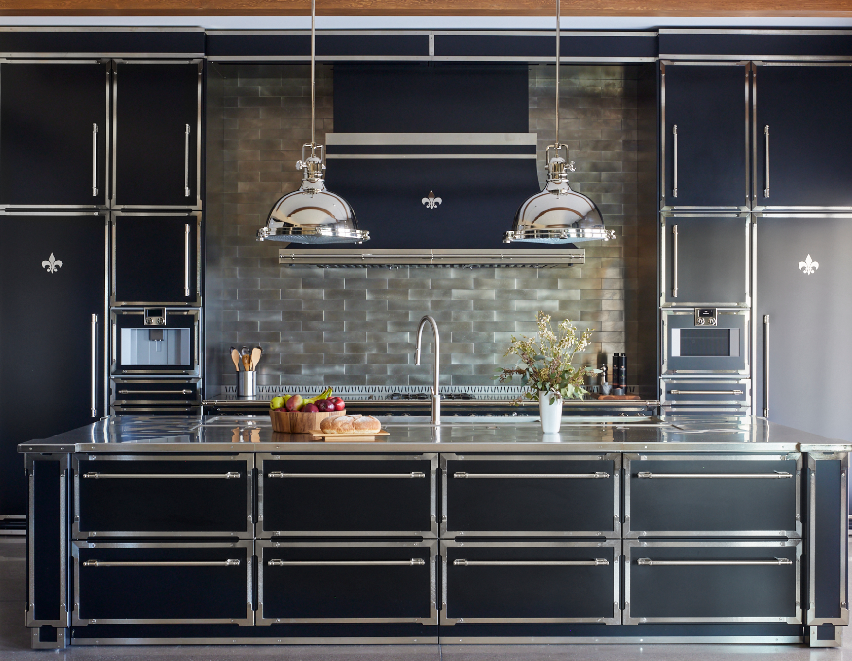 Symmetrical Art Deco-style luxury kitchen with black cabinetry and a large kitchen island, all featuring striking chrome or stainless steel trim and hardware. A custom black range hood with a Fleur de Lis accent is mounted over a metallic subway tile backsplash, illuminated by two large industrial-style chrome pendant lights.
