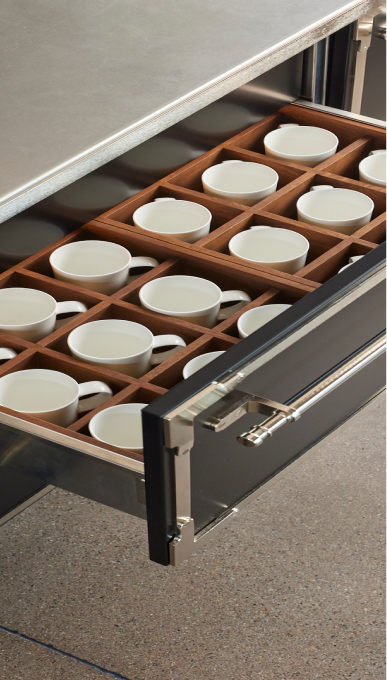 Vertical close-up showing an open black kitchen drawer with chrome trim and handle, revealing a custom wooden organizer insert holding neat rows of white porcelain espresso cups.