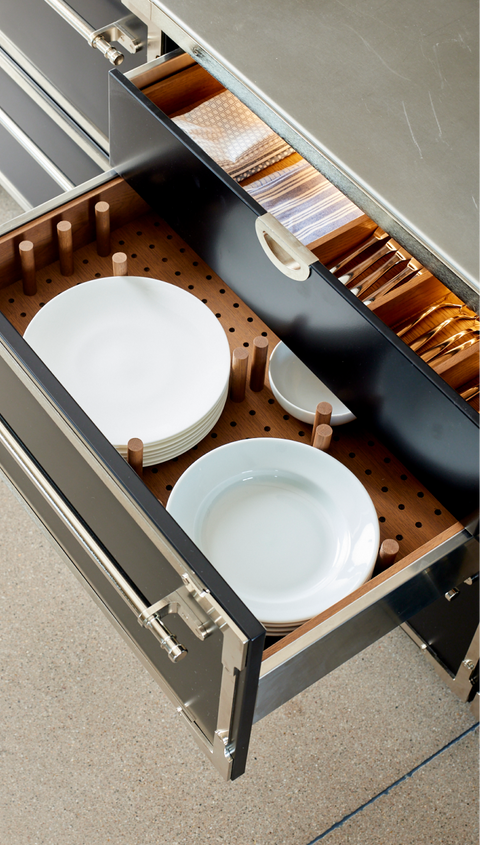 Vertical close-up showing an open black kitchen drawer with chrome trim and handle, featuring a custom wooden plate organizer with removable dowels securely holding stacks of white dinner plates and smaller bowls. A separate upper drawer for silverware is visible above.