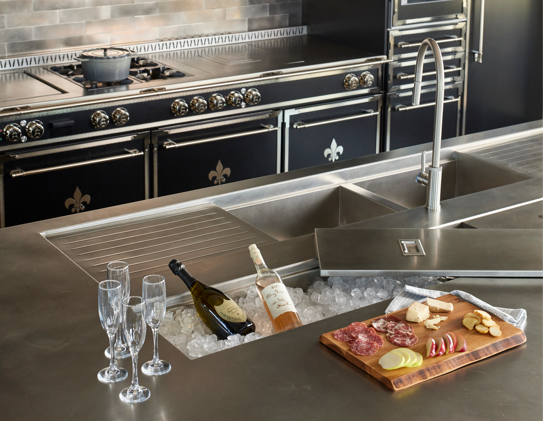 Close-up of a professional stainless steel kitchen island countertop with an integrated sink, drainboard, and a sunken ice well chilling two bottles of wine, with a charcuterie board and champagne flutes nearby. In the background is a black La Cornue range with chrome hardware and a metallic subway tile backsplash.