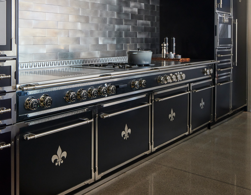 Close-up of a black luxury La Cornue range with chrome trim and stainless steel accents, featuring Fleur de Lis emblems on the oven doors and brass-toned control knobs, set against a metallic gray subway tile backsplash in a modern black kitchen.