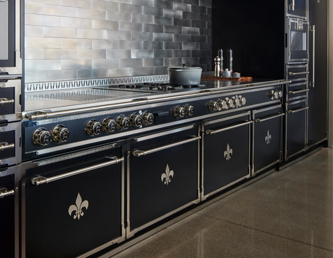 Close-up of a black luxury La Cornue range with chrome trim and stainless steel accents, featuring Fleur de Lis emblems on the oven doors and brass-toned control knobs, set against a metallic gray subway tile backsplash in a modern black kitchen.