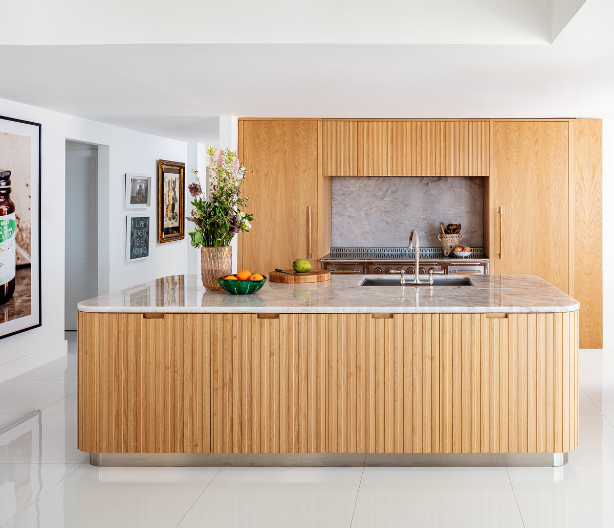 A bright, modern kitchen featuring a large central island with a rounded marble countertop and a striking fluted natural wood base