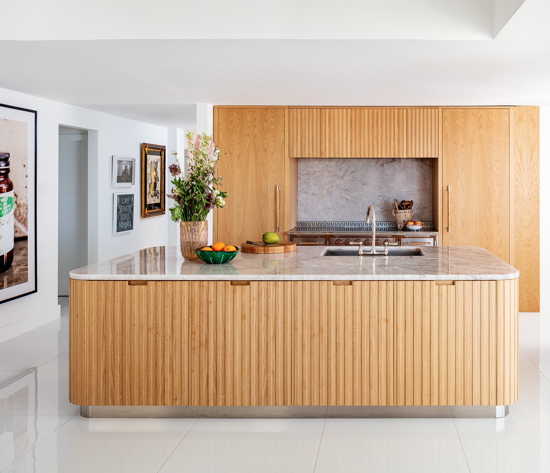 A bright, modern kitchen featuring a large central island with a rounded marble countertop and a striking fluted natural wood base. The island includes an integrated sink with a gooseneck faucet. The back wall features full-height natural wood cabinetry that conceals appliances, with a recess housing a range and a gray marble backsplash. The kitchen has glossy white floor tiles and a vase of flowers on the island.