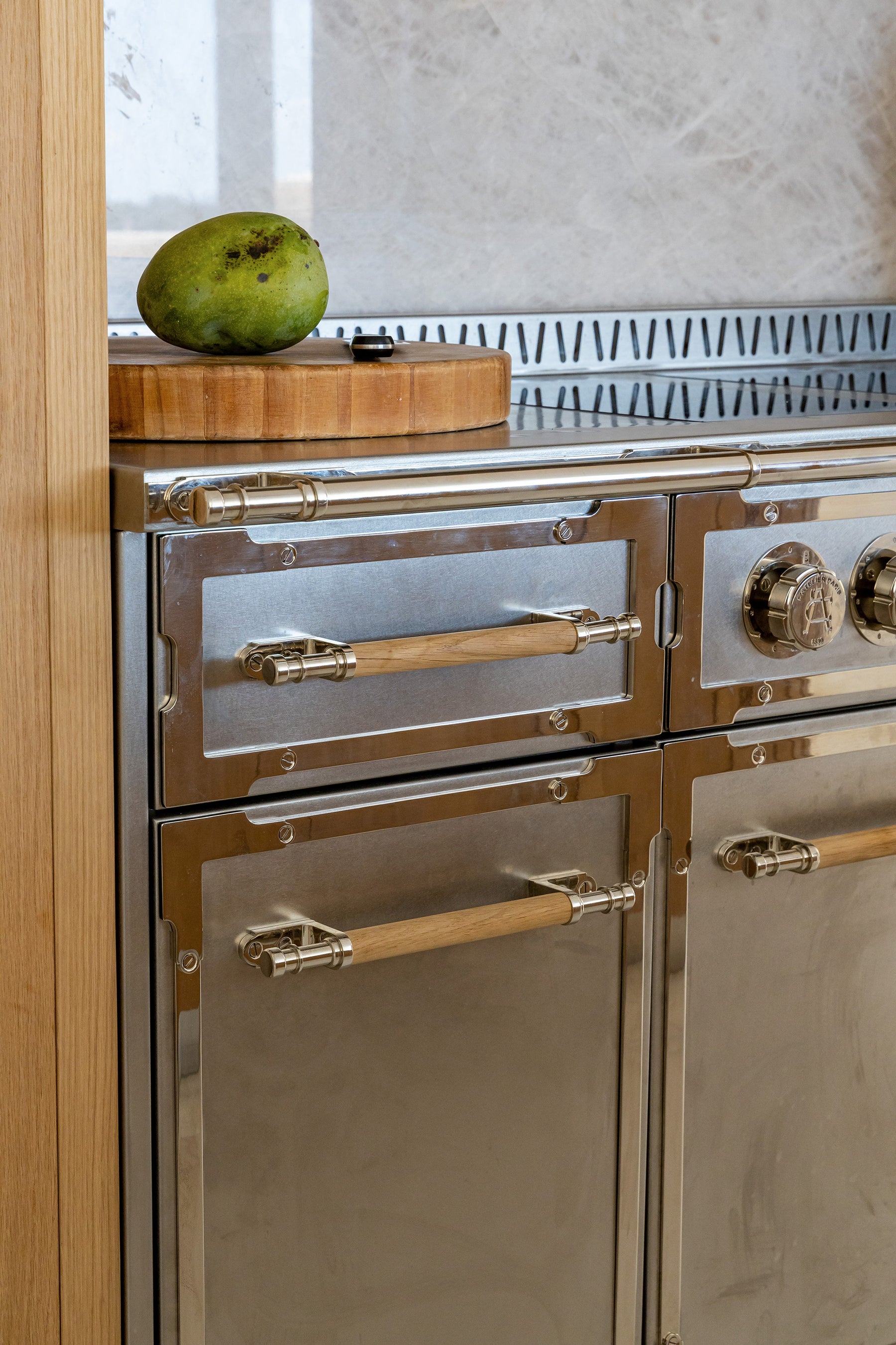 A vertical close-up of a professional stainless steel kitchen range with a brushed finish and polished accents. The oven doors and drawers feature light wooden handles with stylish silver-tone brackets. Above the cooktop, a gray marble slab backsplash is visible next to a wooden cutting board with a green mango.