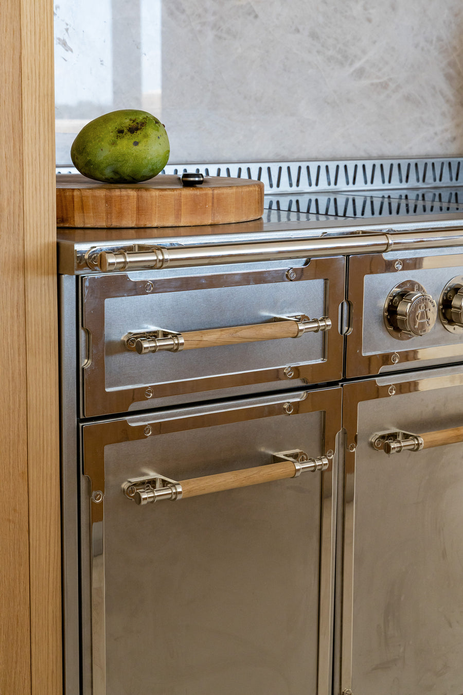 A vertical close-up of a professional stainless steel kitchen range with a brushed finish and polished accents. The oven doors and drawers feature light wooden handles with stylish silver-tone brackets. Above the cooktop, a gray marble slab backsplash is visible next to a wooden cutting board with a green mango.