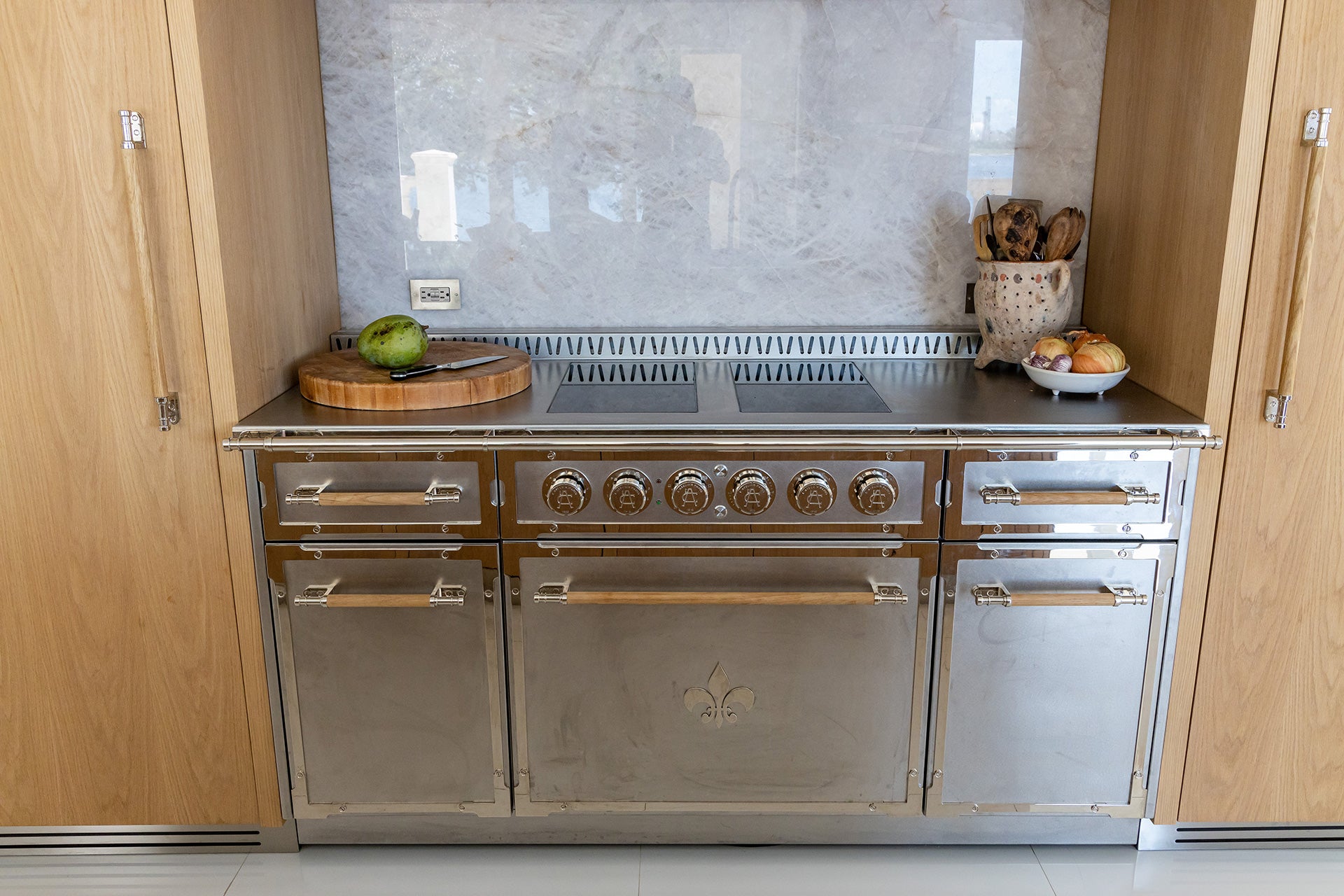 A straight-on shot of a professional stainless steel range with a central Fleur-de-lis emblem and natural wooden handles. The range is seamlessly integrated into a wall of natural wood cabinetry. A stunning gray-veined marble slab backsplash covers the area behind the cooktop, which holds a wooden cutting board and kitchen accessories.