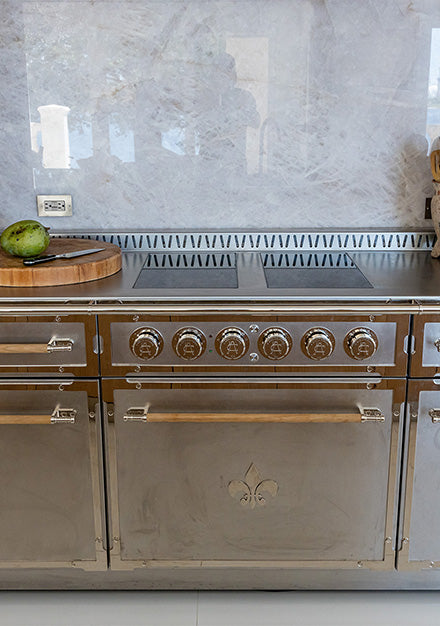 A straight-on shot of a professional stainless steel range with a central Fleur-de-lis emblem and natural wooden handles. The range is seamlessly integrated into a wall of natural wood cabinetry. A stunning gray-veined marble slab backsplash covers the area behind the cooktop, which holds a wooden cutting board and kitchen accessories.