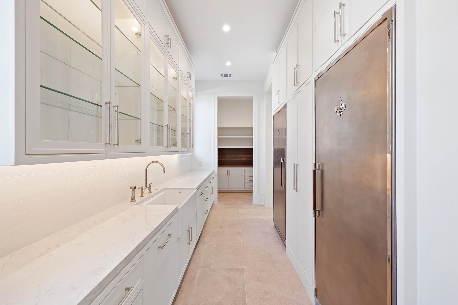 Bright galley kitchen or scullery featuring a long bank of white shaker cabinets with glass-front upper doors and brushed nickel hardware, a white undermount sink, and marble-look countertops. The room has a brushed copper-tone integrated refrigerator, recessed lighting, and leads to a walk-in pantry with wood shelving.