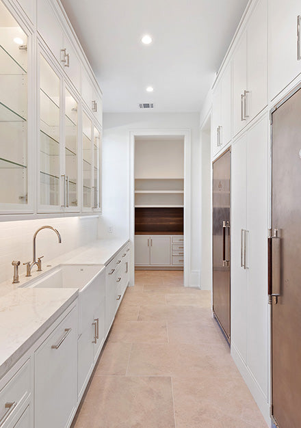 Bright galley kitchen or scullery featuring a long bank of white shaker cabinets with glass-front upper doors and brushed nickel hardware, a white undermount sink, and marble-look countertops. The room has a brushed copper-tone integrated refrigerator, recessed lighting, and leads to a walk-in pantry with wood shelving.
