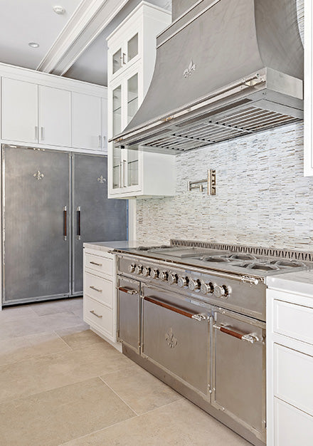 Vertical view of a high-end stainless steel range with wood accents on the handles and Fleur de Lis emblems, under a matching stainless steel hood, set next to a brushed stainless steel paneled refrigerator. The kitchen features white shaker cabinets and a mosaic tile backsplash.