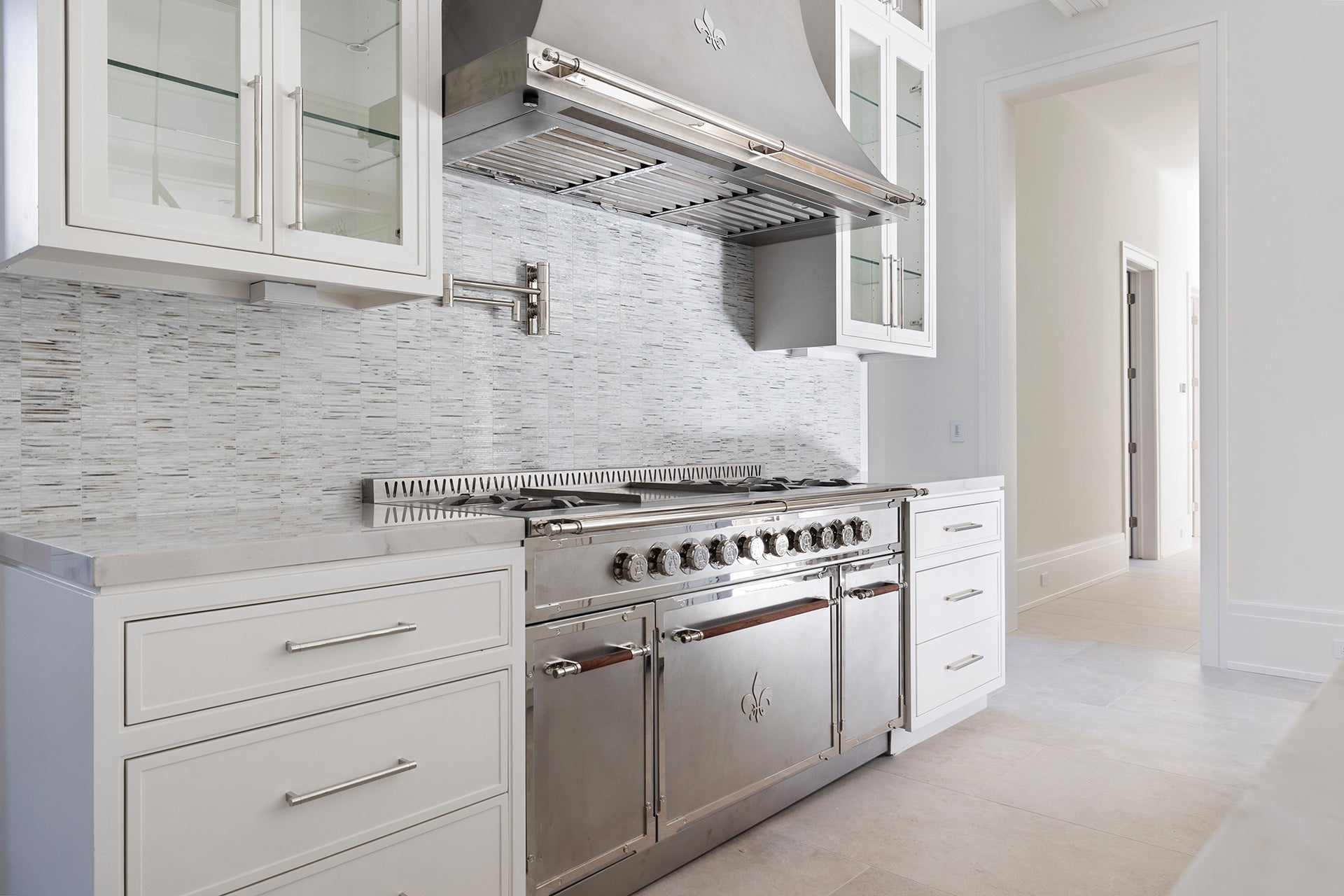 Luxurious contemporary kitchen featuring a large light beige center island with an integrated cooktop and brass trim/hardware, an oversized gold pyramid range hood, dark grey glass-front upper cabinets, and a grey marble slab backsplash, all set on a herringbone wood floor under an ornate ceiling.