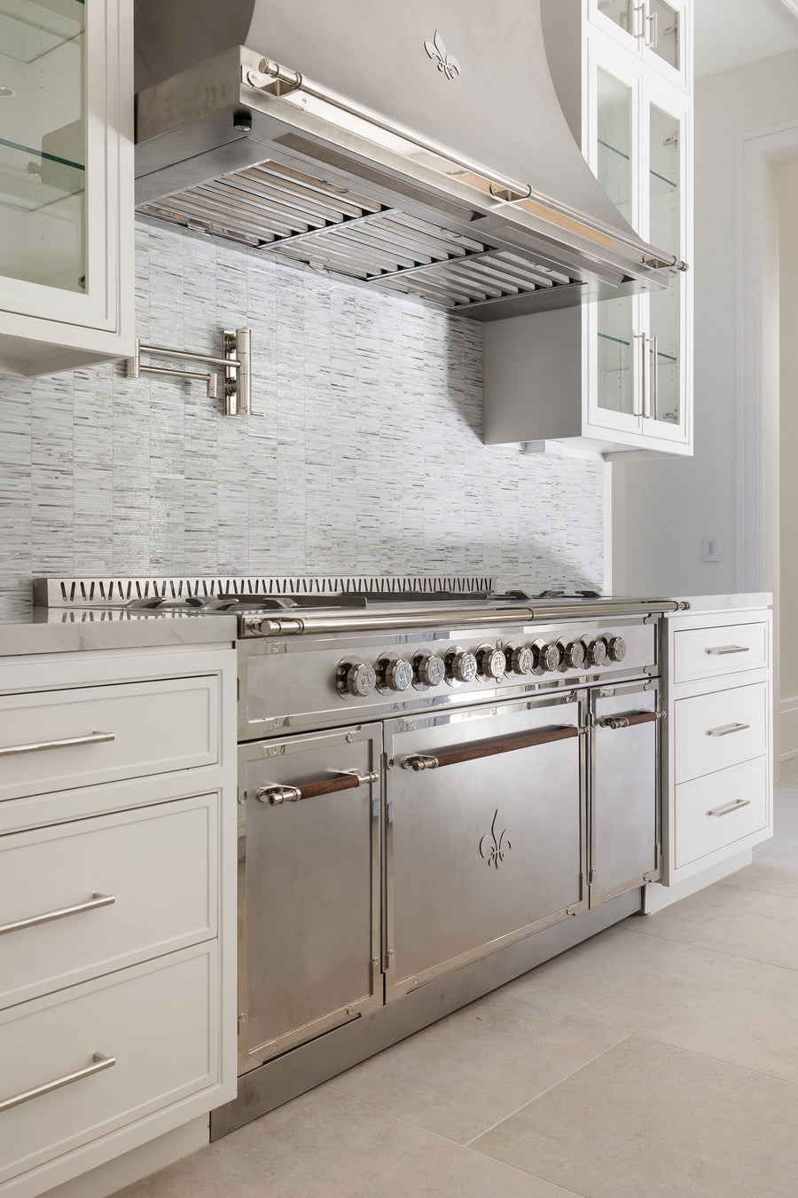 Luxurious contemporary kitchen featuring a large light beige center island with an integrated cooktop and brass trim/hardware, an oversized gold pyramid range hood, dark grey glass-front upper cabinets, and a grey marble slab backsplash, all set on a herringbone wood floor under an ornate ceiling.