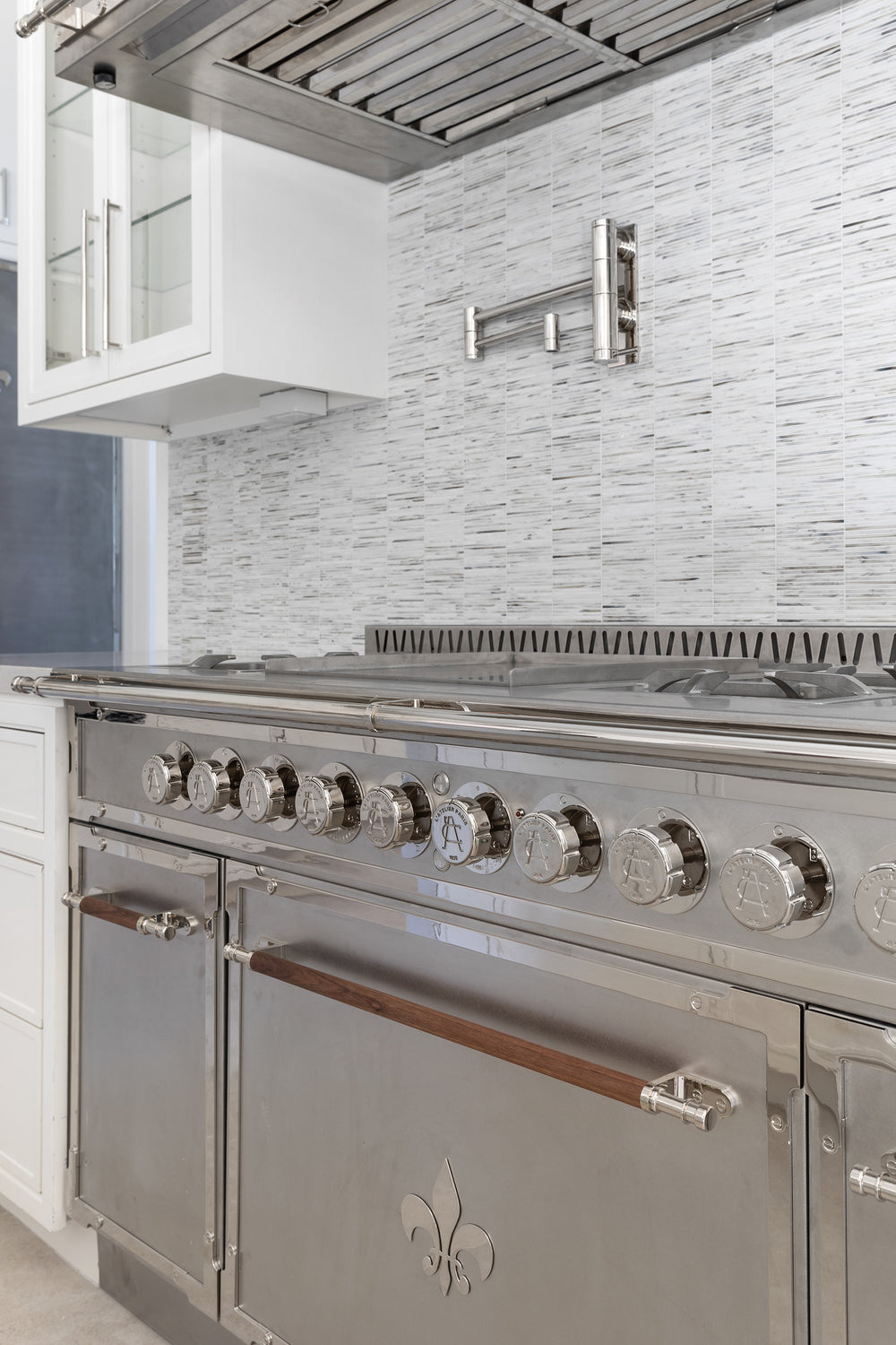 Close-up of a L'Atelier Paris professional stainless steel French range with wood-accented handles and fleur-de-lis detail, featuring a matching chrome pot filler and a textured linear tile backsplash.