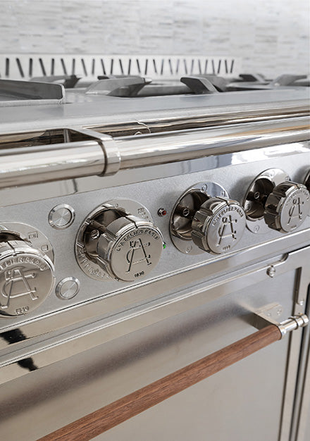 Polished chrome control knobs with L'Atelier Paris 'A. 1830' emblem on a custom stainless steel French kitchen range with wood handle accent.