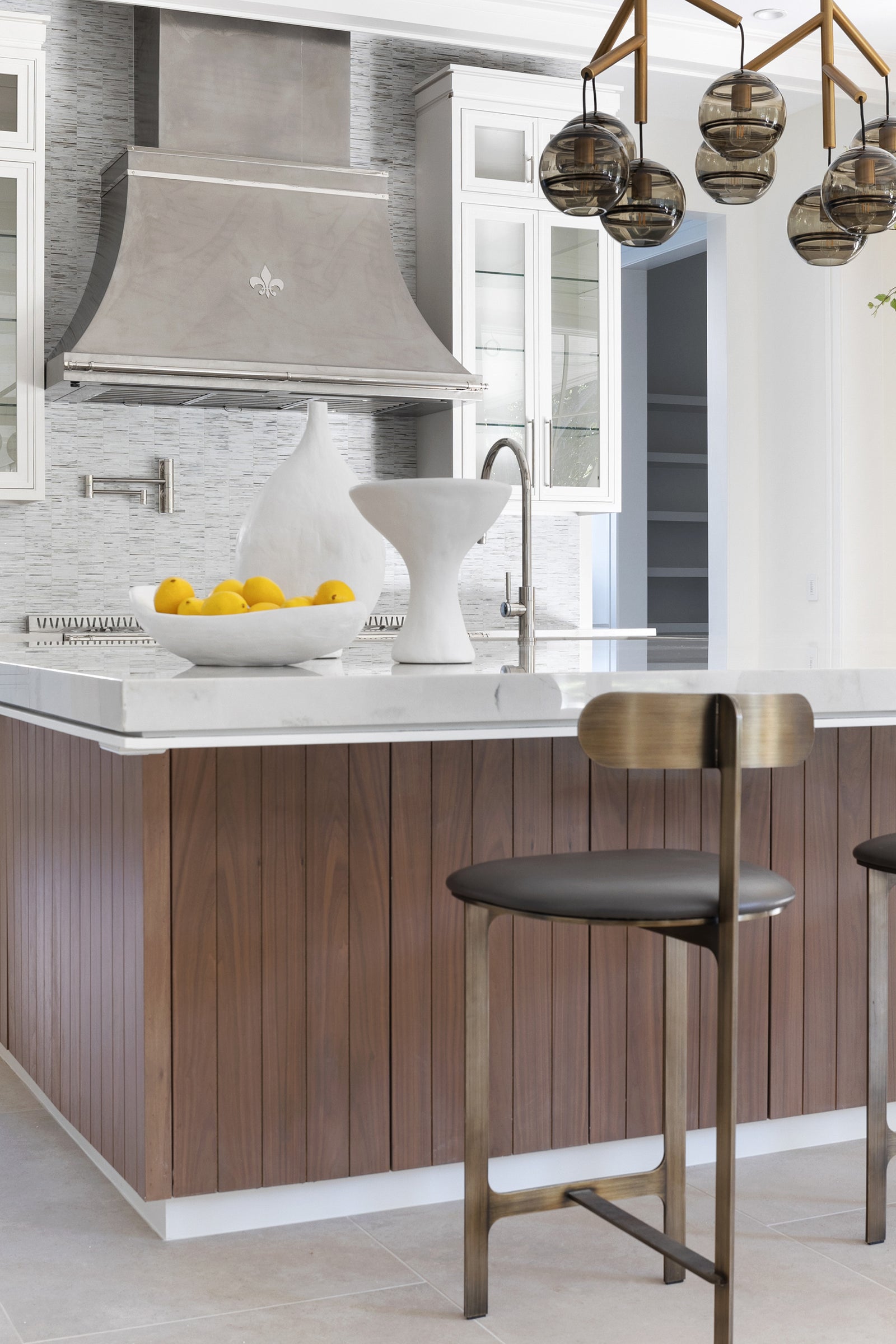 Transitional kitchen design with white marble countertop, walnut slatted island, custom range hood with fleur-de-lis detail, mosaic tile backsplash, and bronze bar stools.