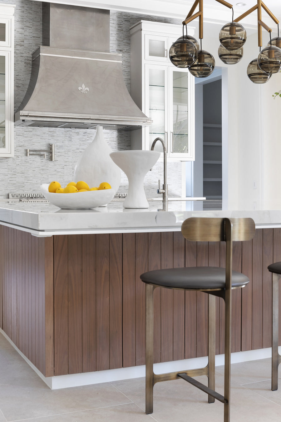 Transitional kitchen design with white marble countertop, walnut slatted island, custom range hood with fleur-de-lis detail, mosaic tile backsplash, and bronze bar stools.
