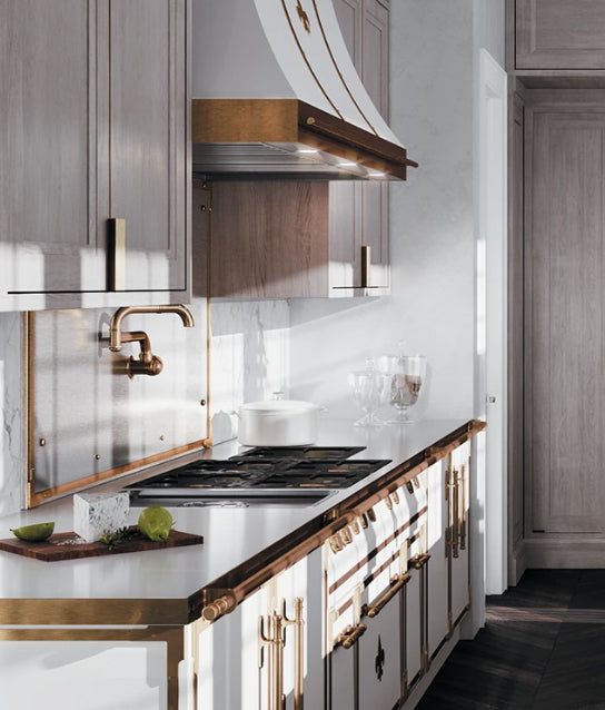 A luxurious and bright kitchen corner featuring a white and bronze professional range with a cooktop and a matching custom range hood. The design includes light gray wood cabinetry, white countertops, a unique bronze pot filler faucet mounted on a metal backsplash, and a dark wood floor.