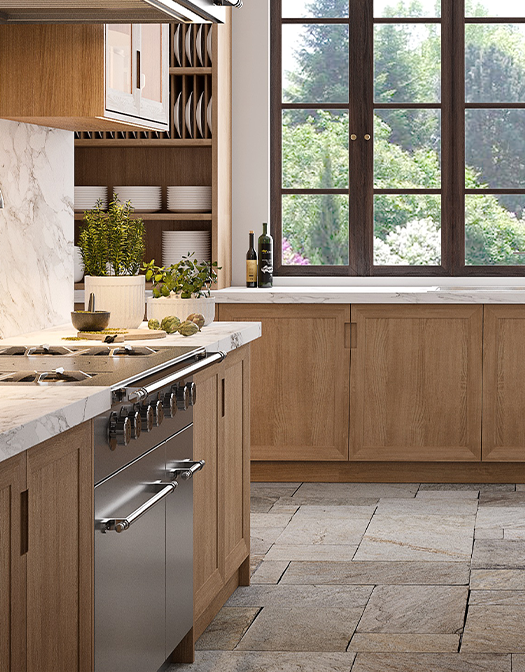 A cozy, transitional kitchen with natural oak cabinetry, a white marble backsplash and countertops, and a stainless steel professional range. The floor is covered in large gray stone tiles, and a prominent, dark-framed window offers a view of a lush green garden.