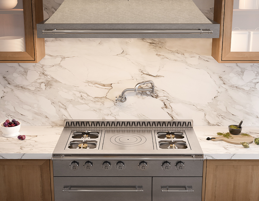 A close-up of a professional stainless steel range with a built-in griddle, set beneath a gray fabric-covered range hood with a silver bar handle. The range is surrounded by white marble backsplash and countertops and natural wood cabinetry, with a pot filler faucet mounted above the cooking surface.