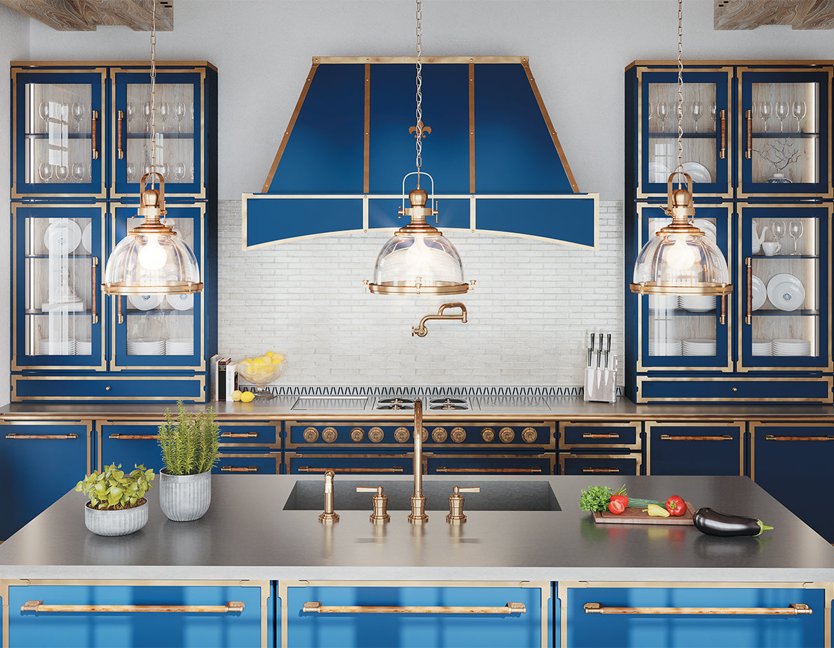 A symmetrical and opulent kitchen featuring bold blue cabinetry with gold trim and hardware, centered around a matching blue professional range and a custom range hood adorned with a fleur-de-lis. The kitchen also has a large island with a dark gray countertop and integrated sink, three industrial-style glass pendant lights, and a white subway tile backsplash with a brass pot filler faucet.
