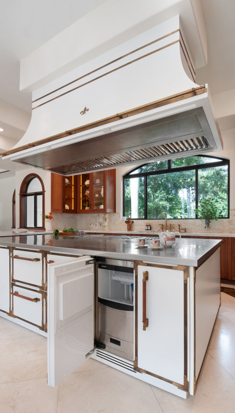 Elegant and spacious kitchen with a large white center island featuring brass accents, a built-in ice machine, and a polished stainless steel countertop. The kitchen includes dark wood cabinetry, a decorative range hood with a unique emblem, and large arched windows with a view of the outdoors.