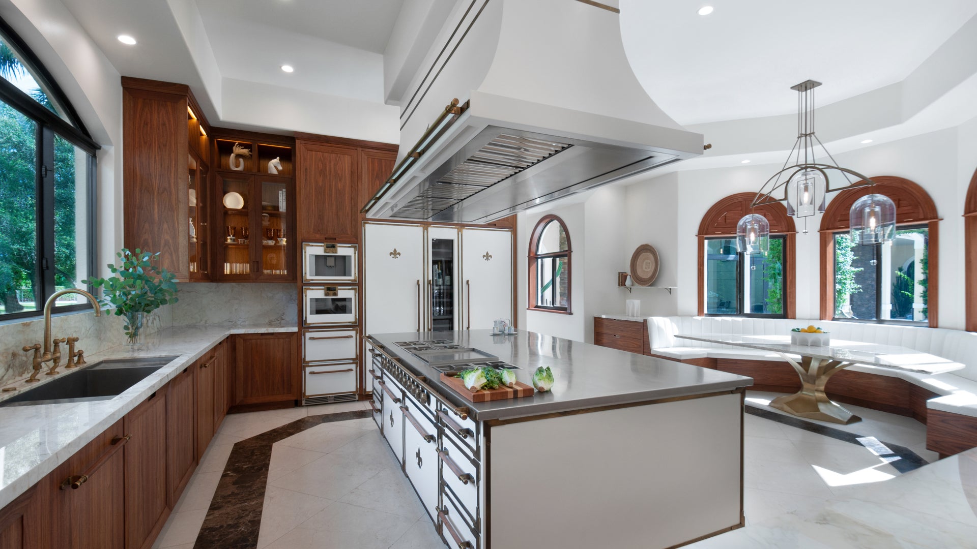 A magnificent French-style kitchen with a large, central island featuring a stainless steel top and an integrated cooktop. The kitchen boasts luxurious dark wood cabinetry, white marble countertops, and a prominent white and brass range hood. Integrated refrigerators with white doors and fleur-de-lis emblems are visible, and the space includes a cozy built-in banquette seating area with arched windows.