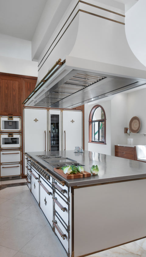 A high-angle view of a luxurious French-style kitchen island with a stainless steel countertop and integrated cooktop. Above it, a grand white and brass range hood dominates the scene, while built-in white refrigerators with fleur-de-lis emblems and dark wood cabinetry line the background.