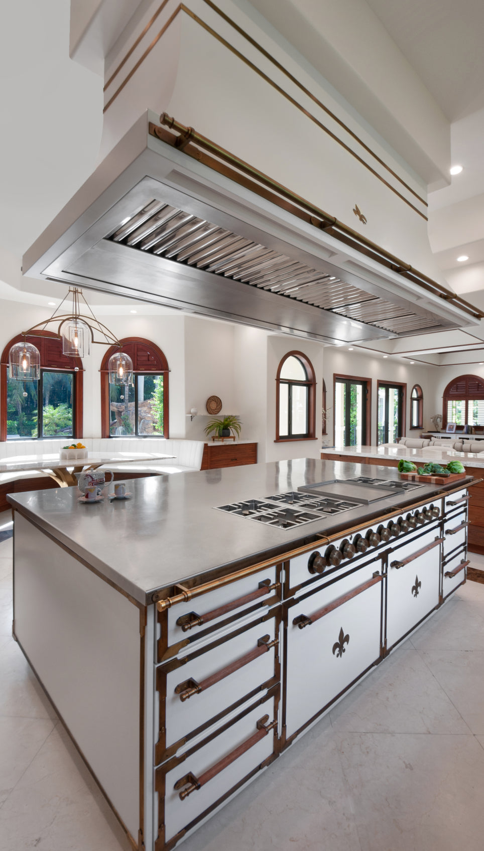 Elegant and spacious kitchen with a large white center island that has a built-in stovetop, oven, and drawers with unique brass and wood handles. A decorative range hood hangs overhead, and a breakfast nook with arched windows is visible in the background.
