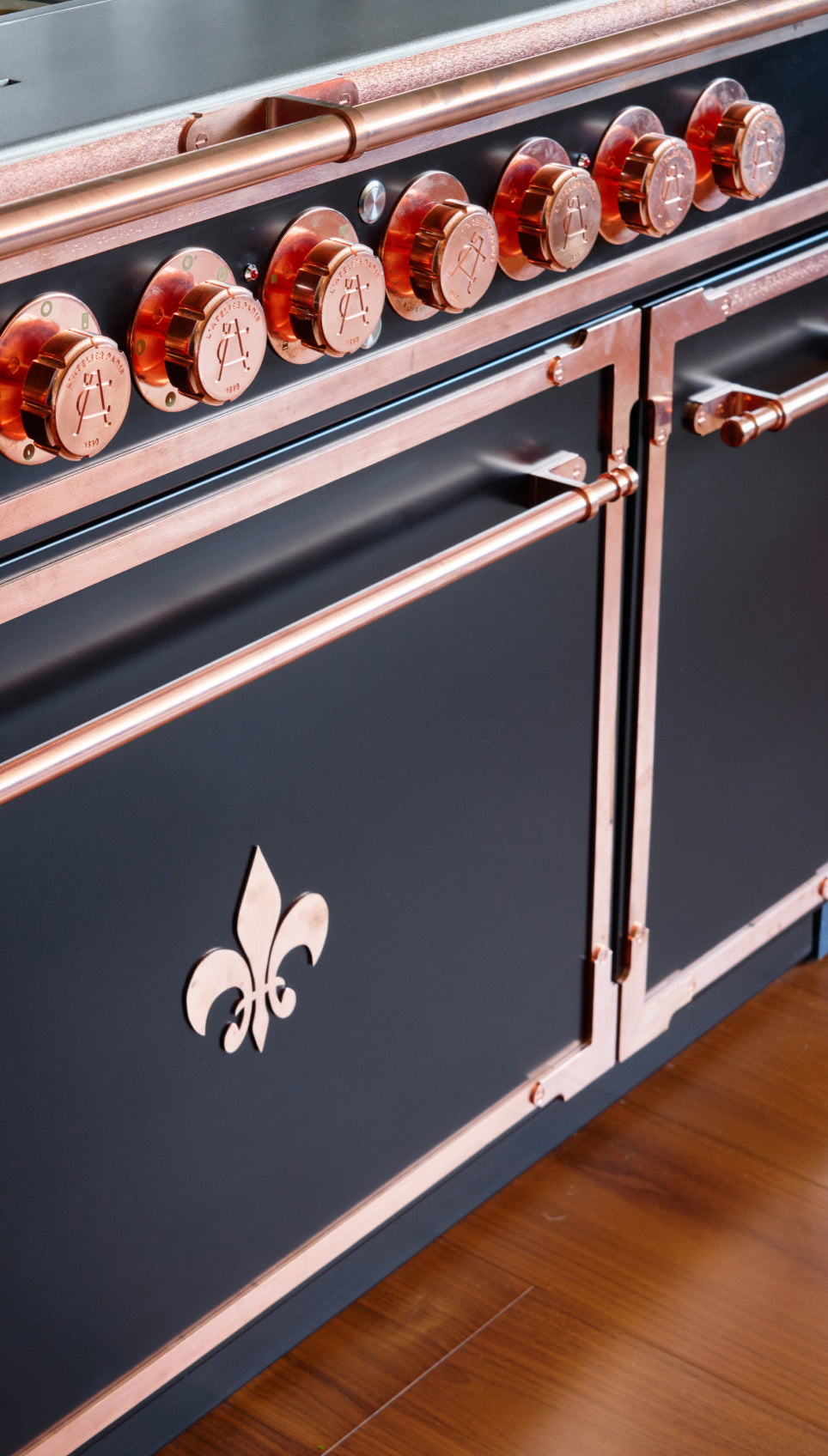 Close-up of a luxury French kitchen range with copper knobs and black matte finish featuring a fleur-de-lis emblem.