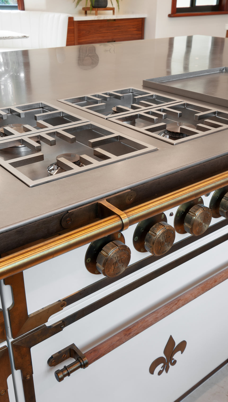 A close-up of a high-end French kitchen range with a stainless steel cooktop and a brass handle bar. The front of the oven is white with a fleur-de-lis emblem and large, industrial-style knobs.