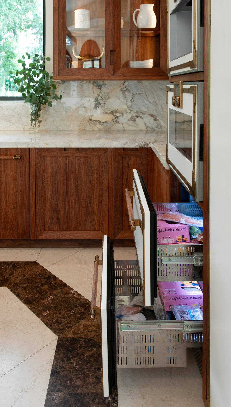 A custom kitchen featuring an integrated freezer drawer pulled out, revealing its contents. The space showcases rich dark wood cabinetry, a white marble countertop with brown veining, and unique geometric floor tiles in brown and white, all accented by elegant brass hardware.
