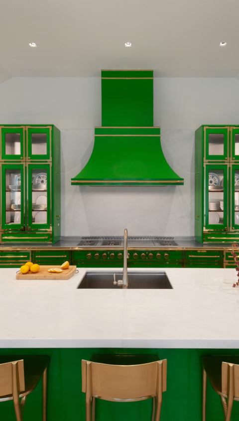 A stylish green kitchen featuring a custom green range hood that matches the glossy cabinets with gold trim. A white island with a built-in sink and a cutting board is in the foreground, with glass display cabinets on either side of the stovetop.