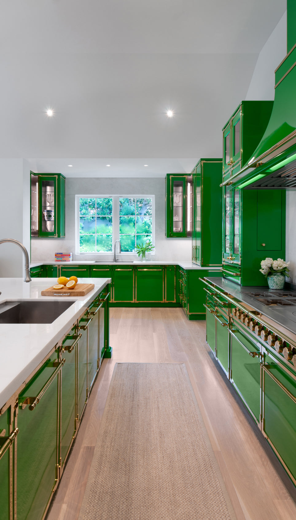 Long view of a luxurious galley-style kitchen featuring glossy green cabinets and gold trim. The space includes a white island with a sink in the foreground, a large window above another sink in the back, and light wood flooring.
