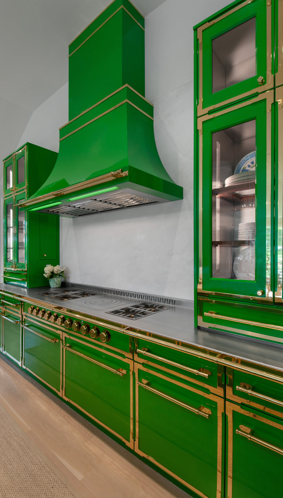 Stylish kitchen featuring glossy emerald green cabinets with gold trim and hardware. A prominent green range hood hangs over a large stainless steel stovetop, flanked by glass display cabinets filled with dishes.